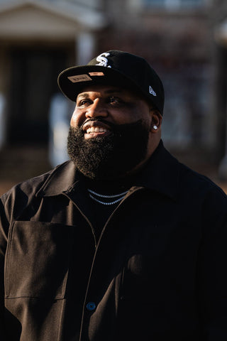 Man with full beard wearing a black cap and dark jacket with a blurred background